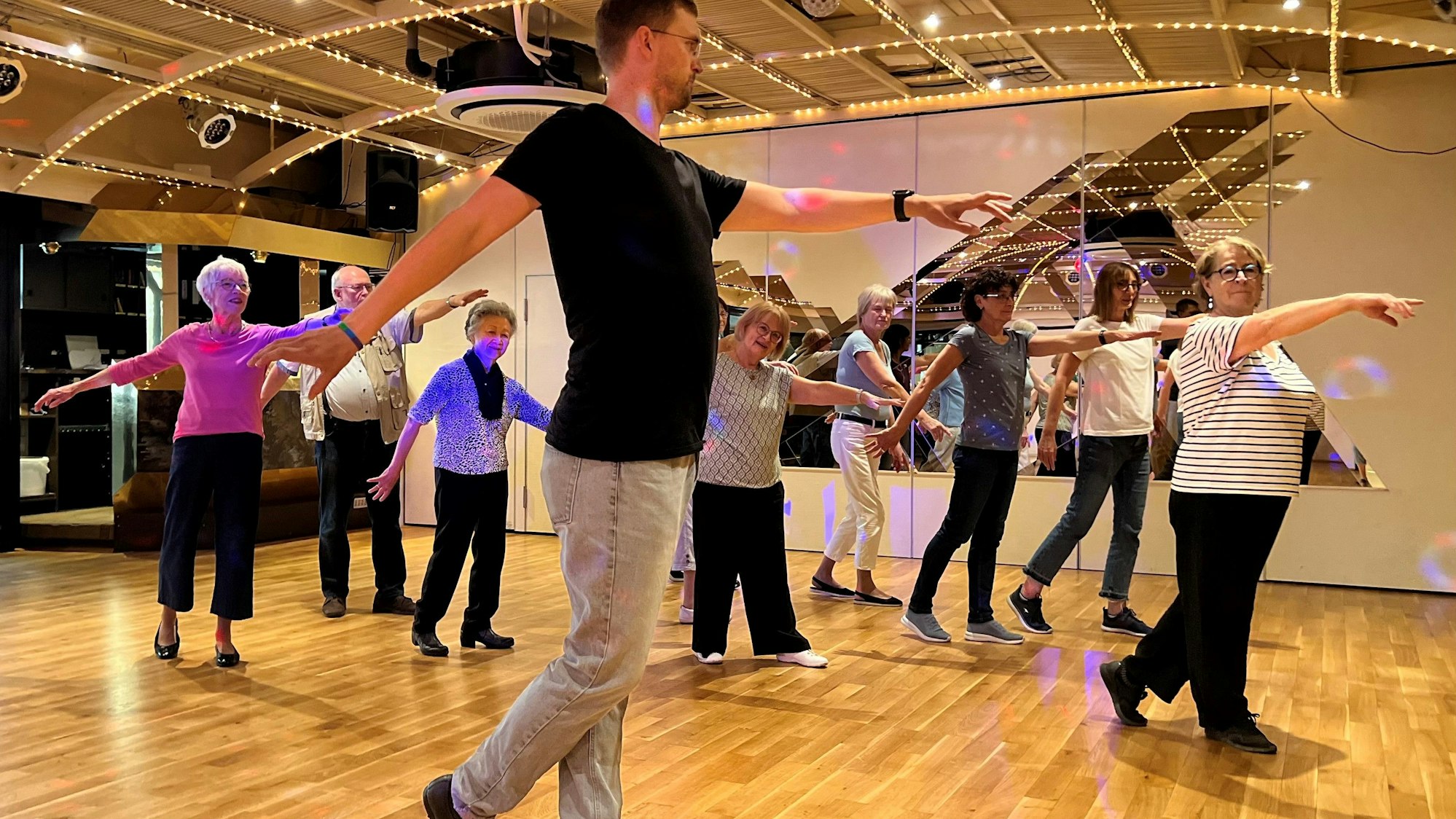 Ältere Damen bewegen sich mit ausgebreiteten Armen über das Parkett, im Vordergrund macht ein jüngerer Mann die Bewegung vor.