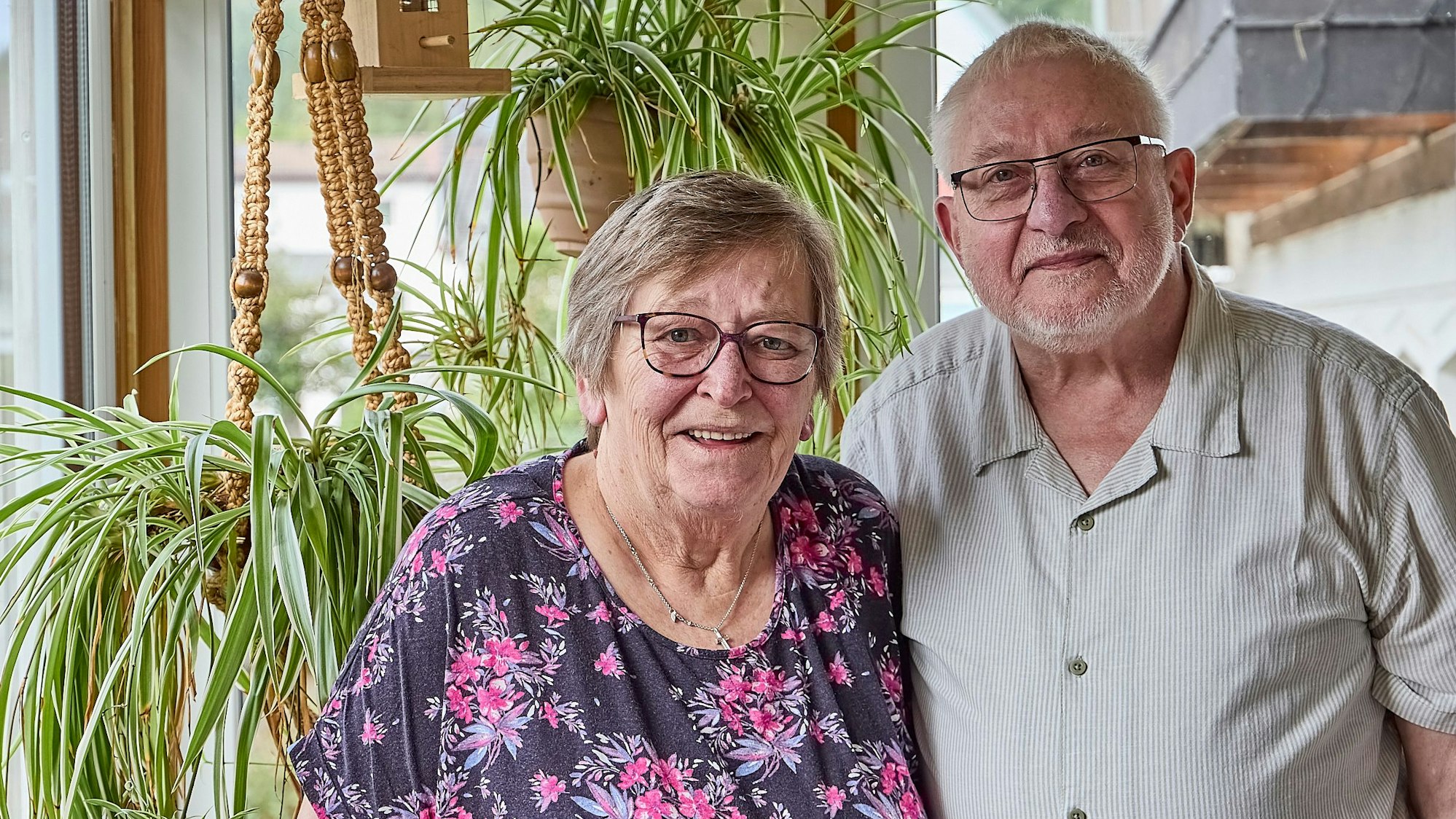 Betty und Hans Herzet stehen nah nebeneinander in einer Wohnung. Sie tragen beide Brille und lächeln in die Kamera. Im Hintergrund befinden sich Pflanzen.