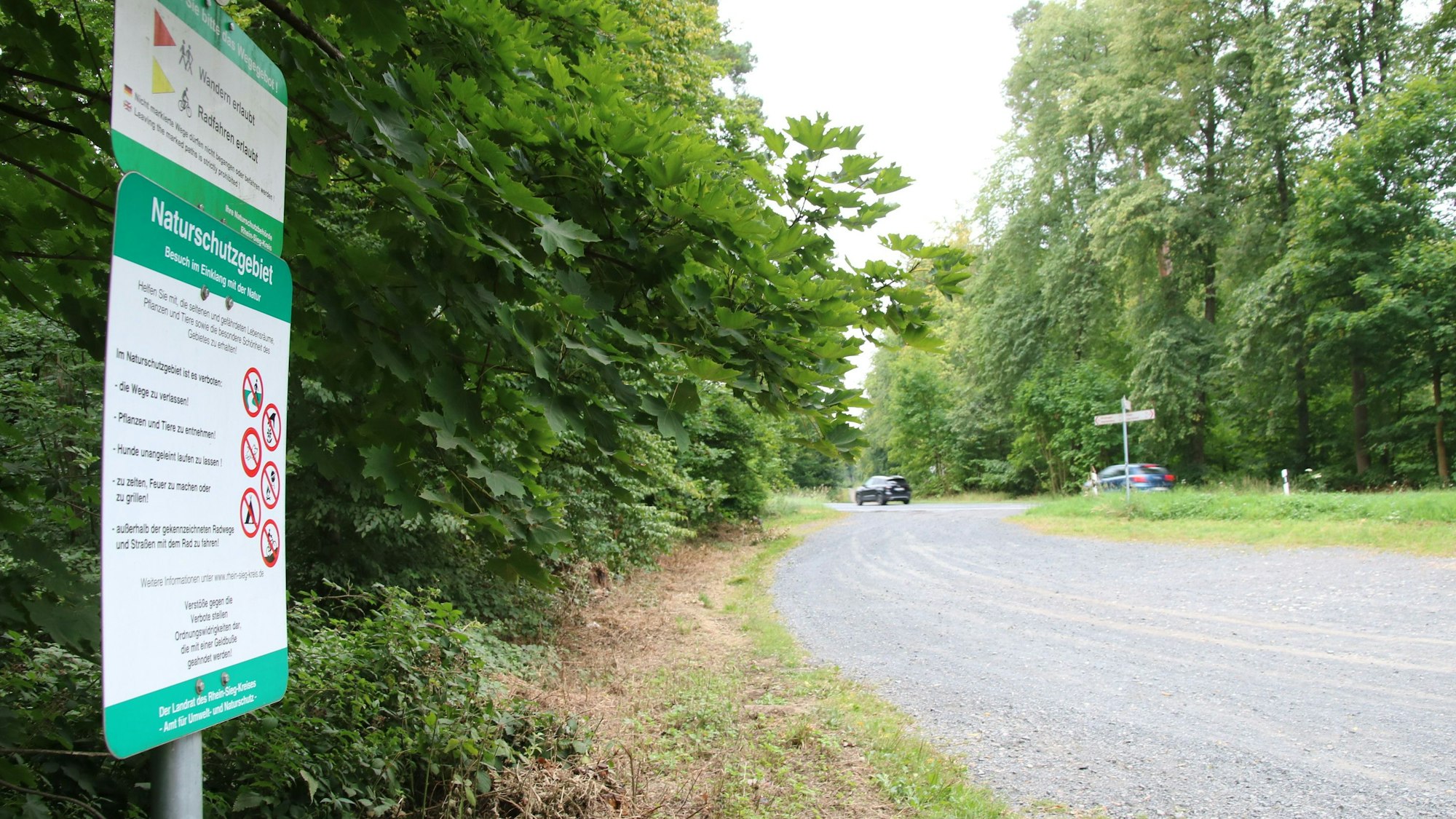 Ein Schild mit der Aufschrift Naturschutzgebiet am Rande eines Parkplatzes; im Hintergrund fahren Autos über eine Straße.