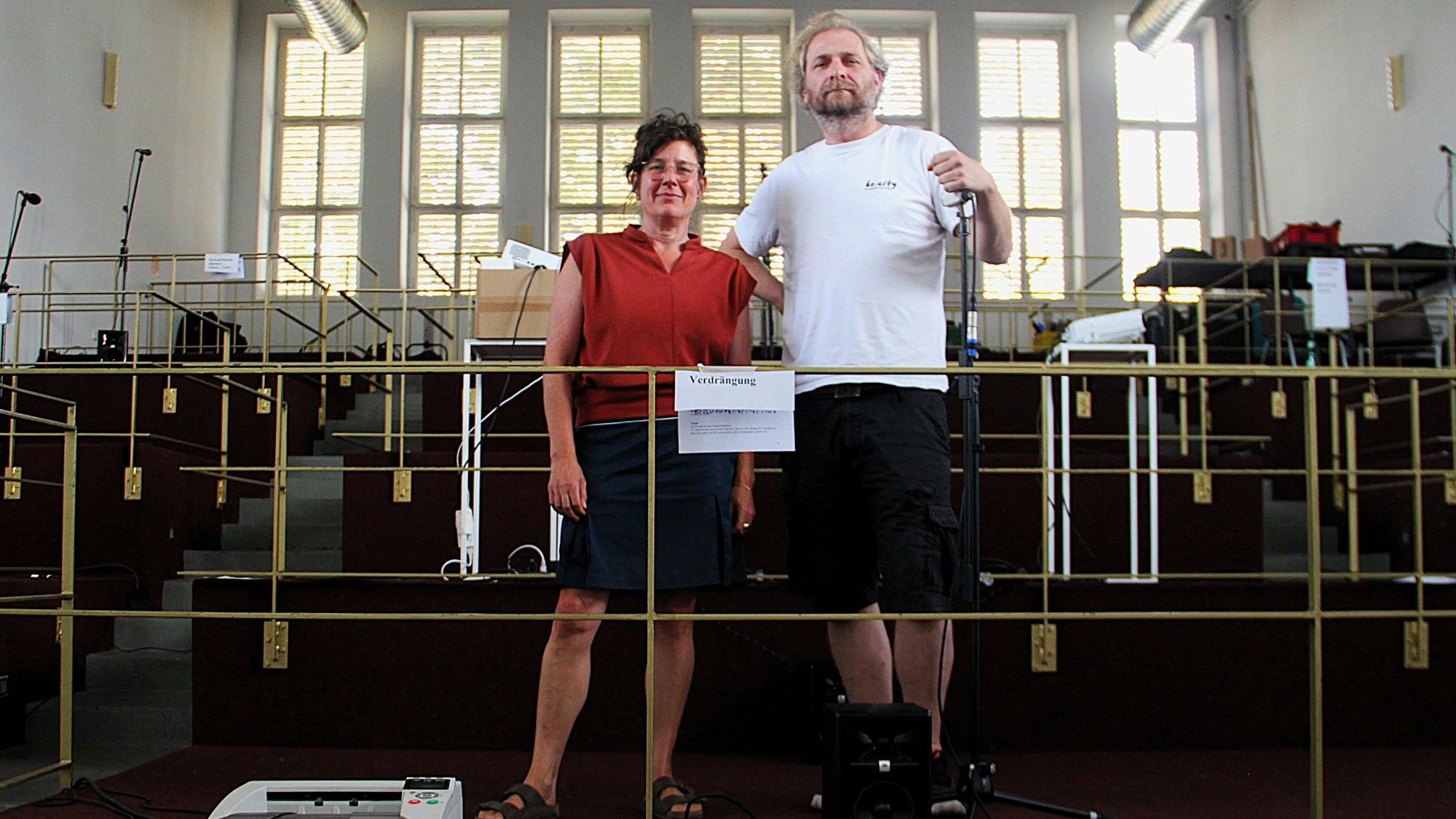 Eine Frau und ein Mann stehen auf einer Art Tribüne in einer großen Halle.