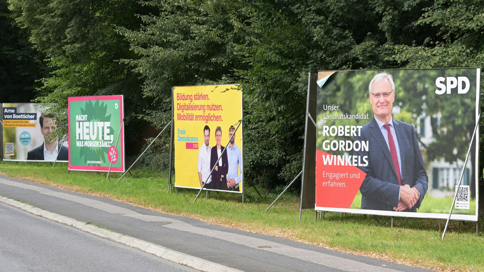 Plakate der CDU, Grünen, FDP und SPD stehen nebeneinander auf der Overather Straße.