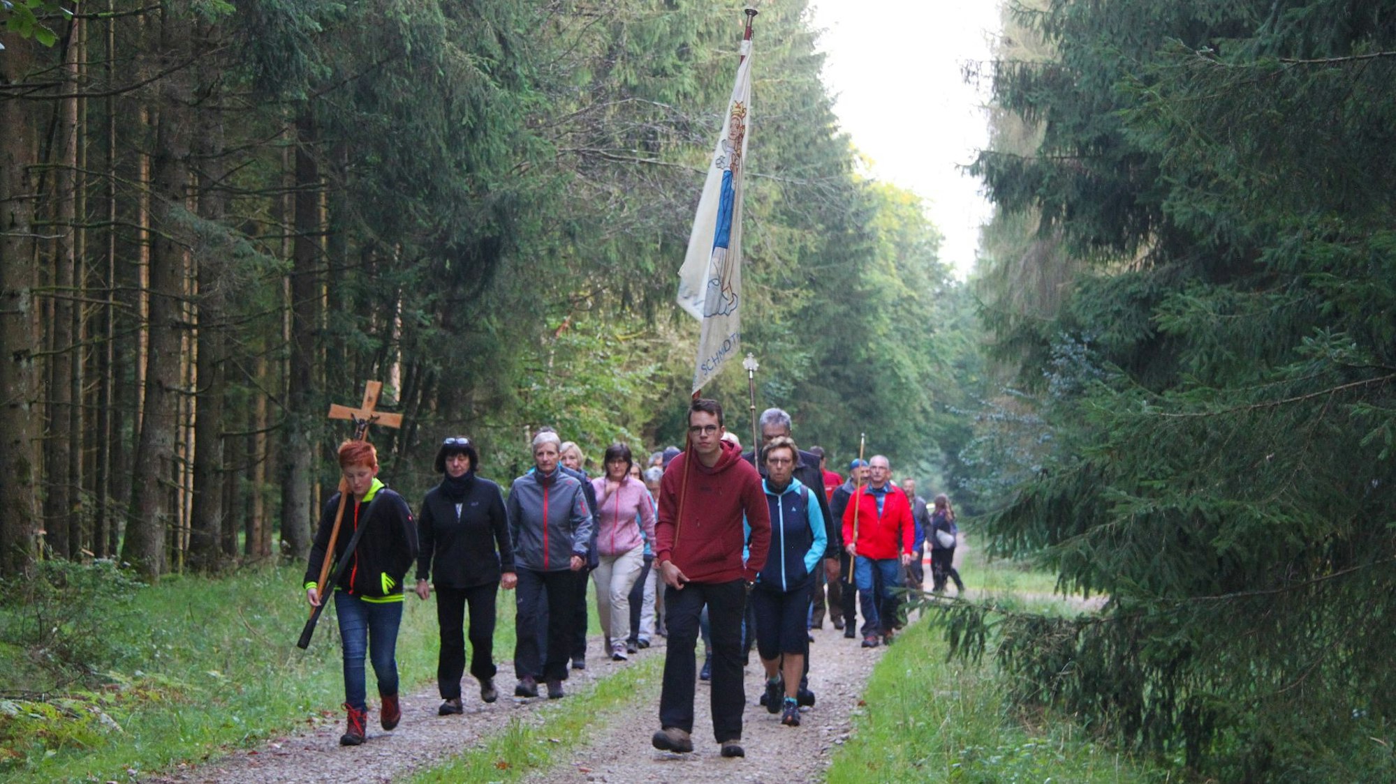 Etwa 20 Personen, die durch einen Wald gehen, sind auf dem Bild zu sehen. Eine Person trägt ein Kreuz, eine andere eine Fahne.