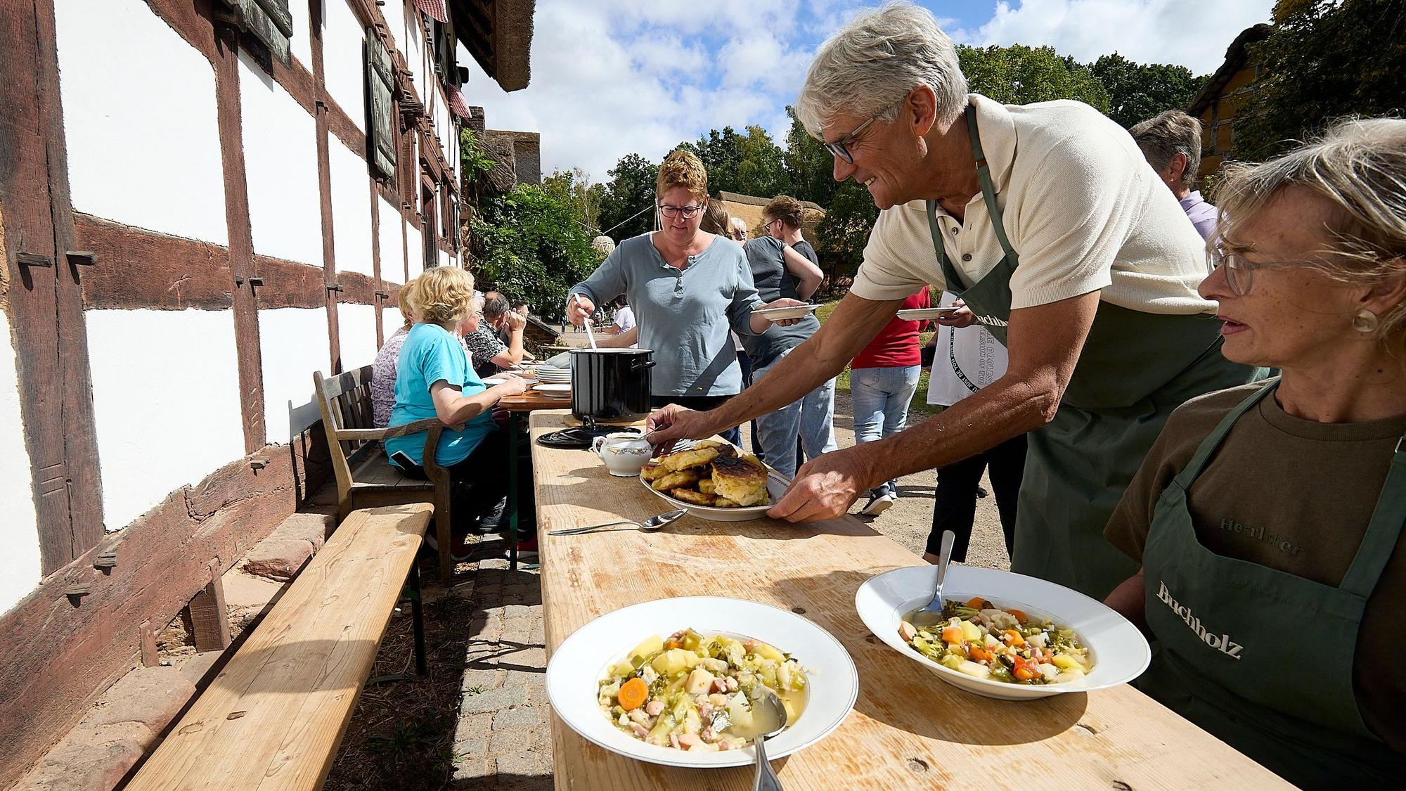 Eine Frau sitzt an einem Tisch vor einem alten Fachwerkhaus, auf dem zwei Teller Suppe stehen. Ein Mann stellt einen Teller mit Pfannkuchen dazu. Im Hintergrund schöpft eine Frau Suppe aus einem großen Kessel.