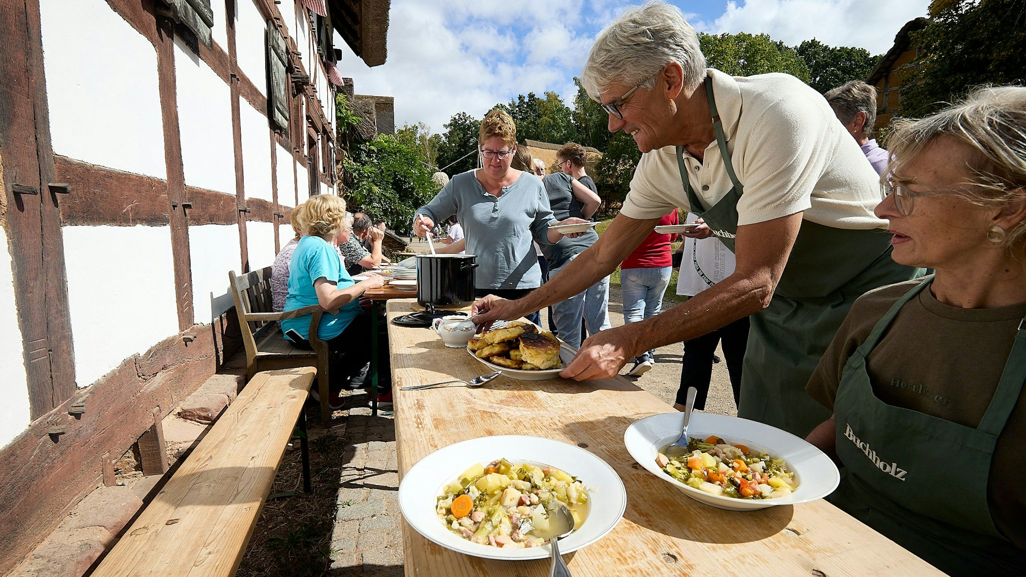 Eine Frau sitzt an einem Tisch vor einem alten Fachwerkhaus, auf dem zwei Teller Suppe stehen. Ein Mann stellt einen Teller mit Pfannkuchen dazu. Im Hintergrund schöpft eine Frau Suppe aus einem großen Kessel.