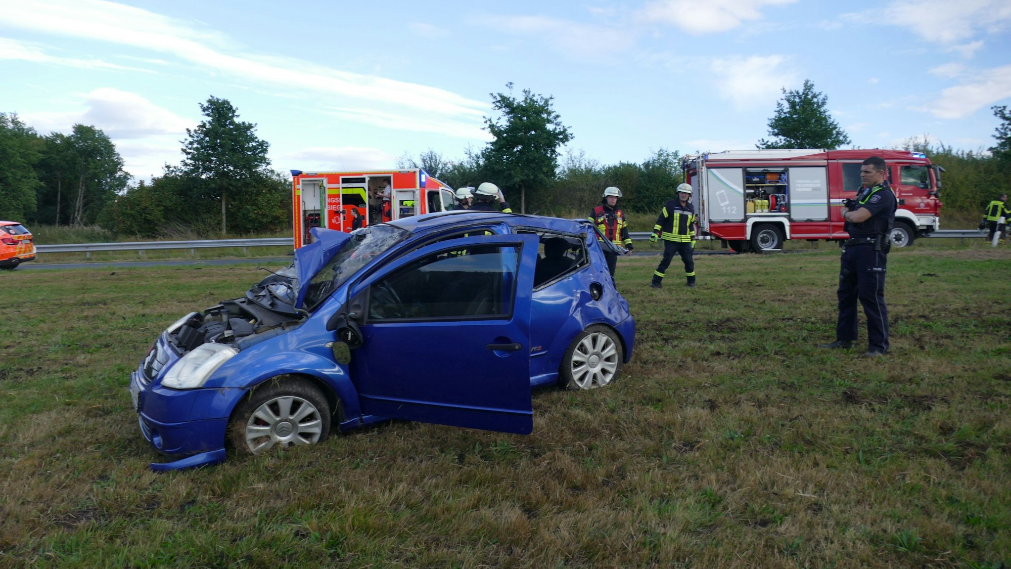 Ein blaues Auto steht auf einer Wiese. Die Fahrertür ist geöffnet, an der Motorhaube und am Heck ist das Auto stark beschädigt.