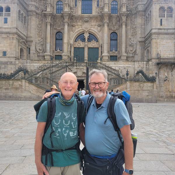 Zwei Männer mit Wandergepäck stehen Arm in Arm vor einer Kirche.