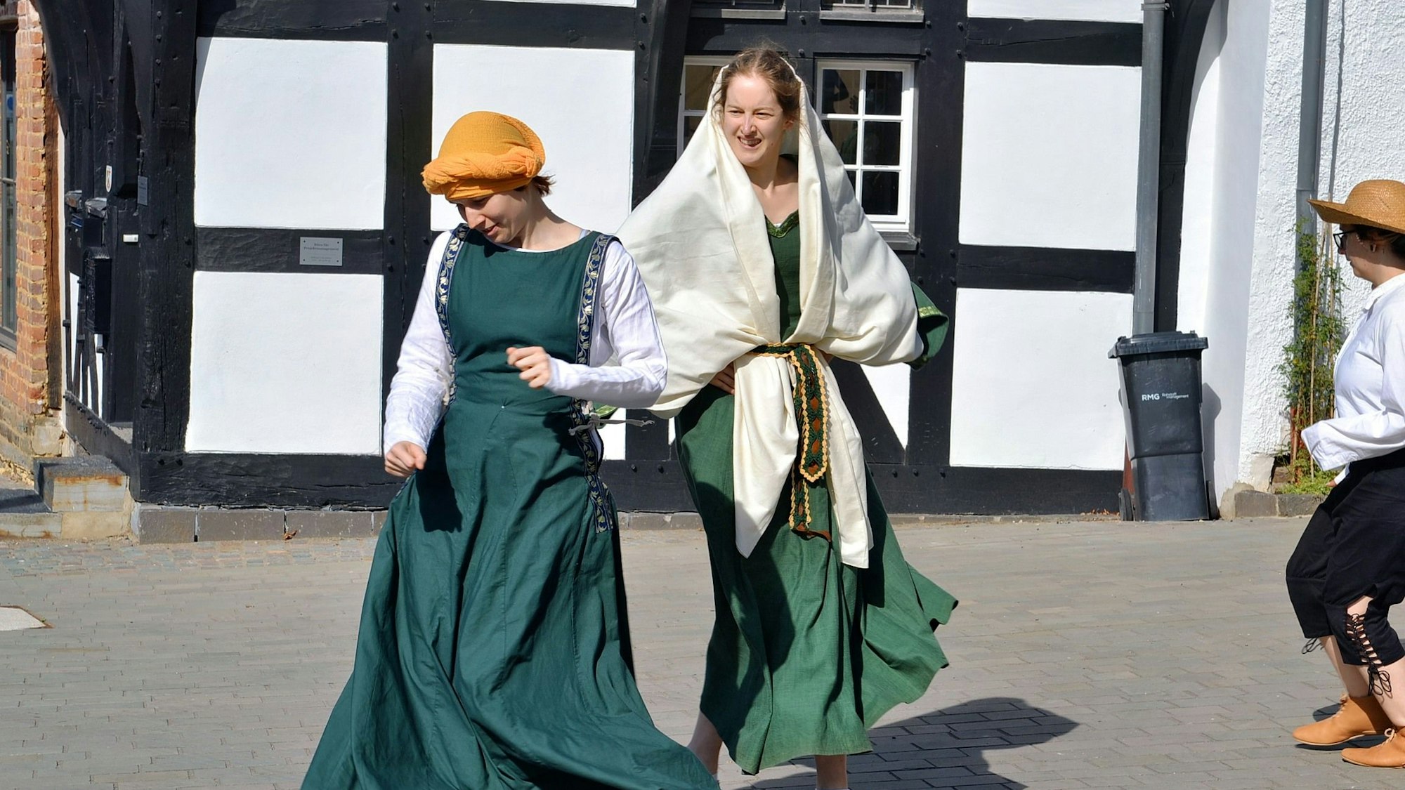 Zwei verkleidete Frauen in Gewändern tanzen vor einem Fachwerkhaus. Im Hintergrund tanzt eine weitere Schauspielerin, die Hut und Brille trägt.