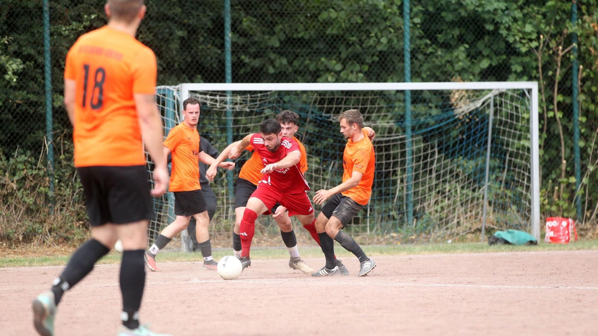 Ein Spieler in einem roten Dress kann den Ball gegen zwei Spieler in orangefarbenem Trikot und schwarzer Hose behaupten.