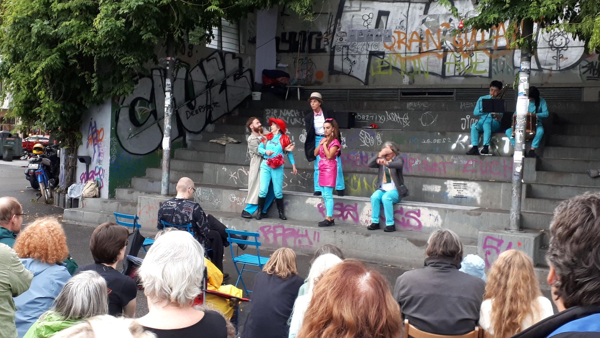 Draußen, auf grauen Treppenstufen, führt ein Ensemble ein Theaterstück auf. Vorne sitzen Zuschauer.