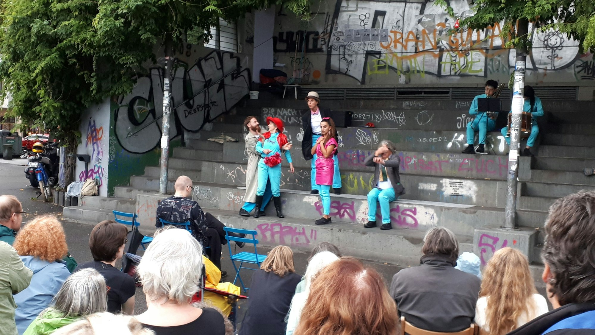 Draußen, auf grauen Treppenstufen, führt ein Ensemble ein Theaterstück auf. Vorne sitzen Zuschauer.