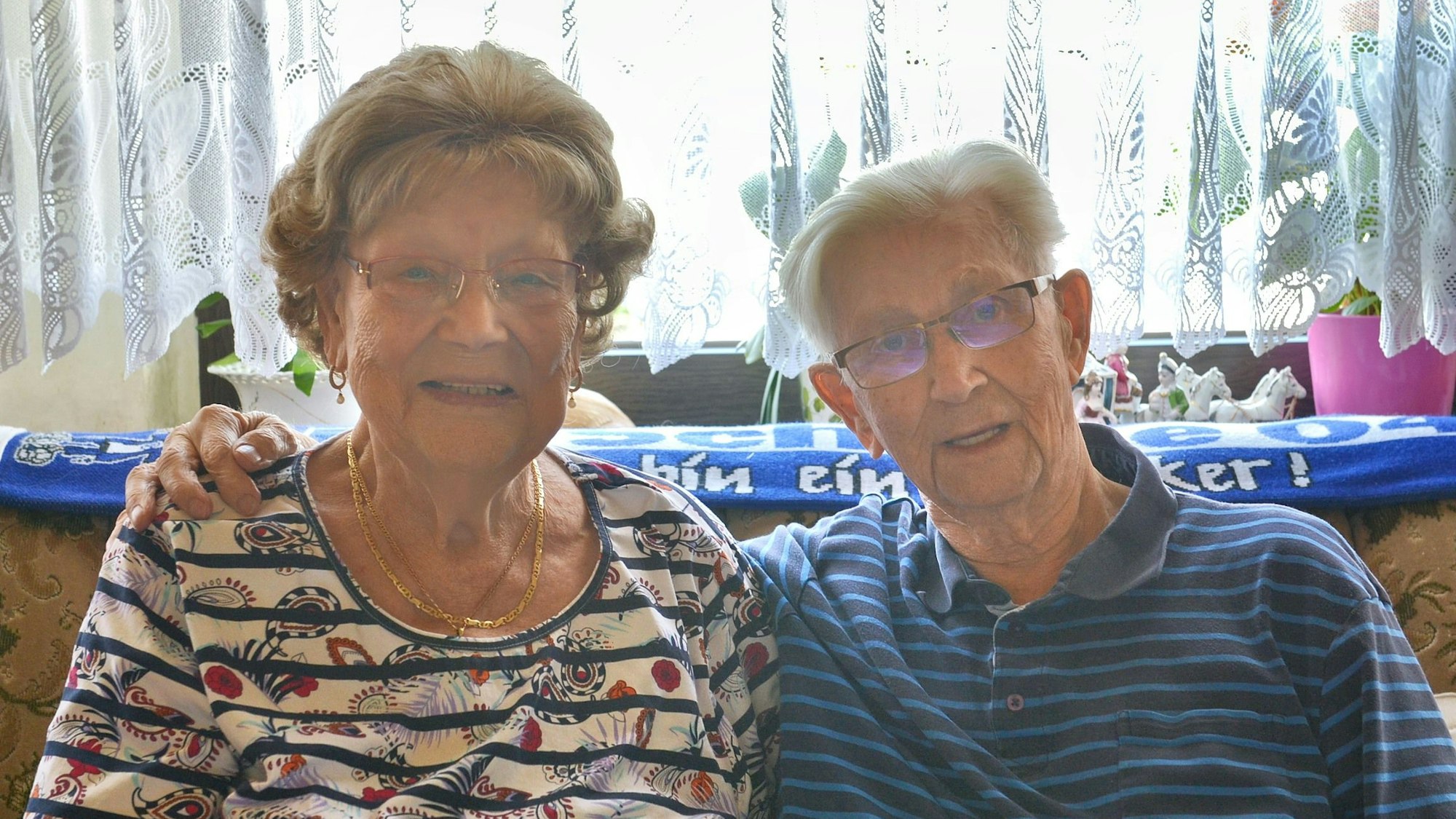 Gertrud Elisabeth und Rudolf Pohl sitzen vor einem hellen Vorhang, durch den Licht scheint. Er hält seinen rechten Arm um sie.