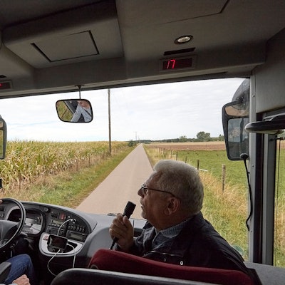 Ein Bus fährt über einen Wirtschaftsweg an einem Maisfeld entlang. Neben dem Fahrer sitzt der Bürvenicher Ortsvorsteher Jörg Körtgen und spricht in ein Mikrofon.