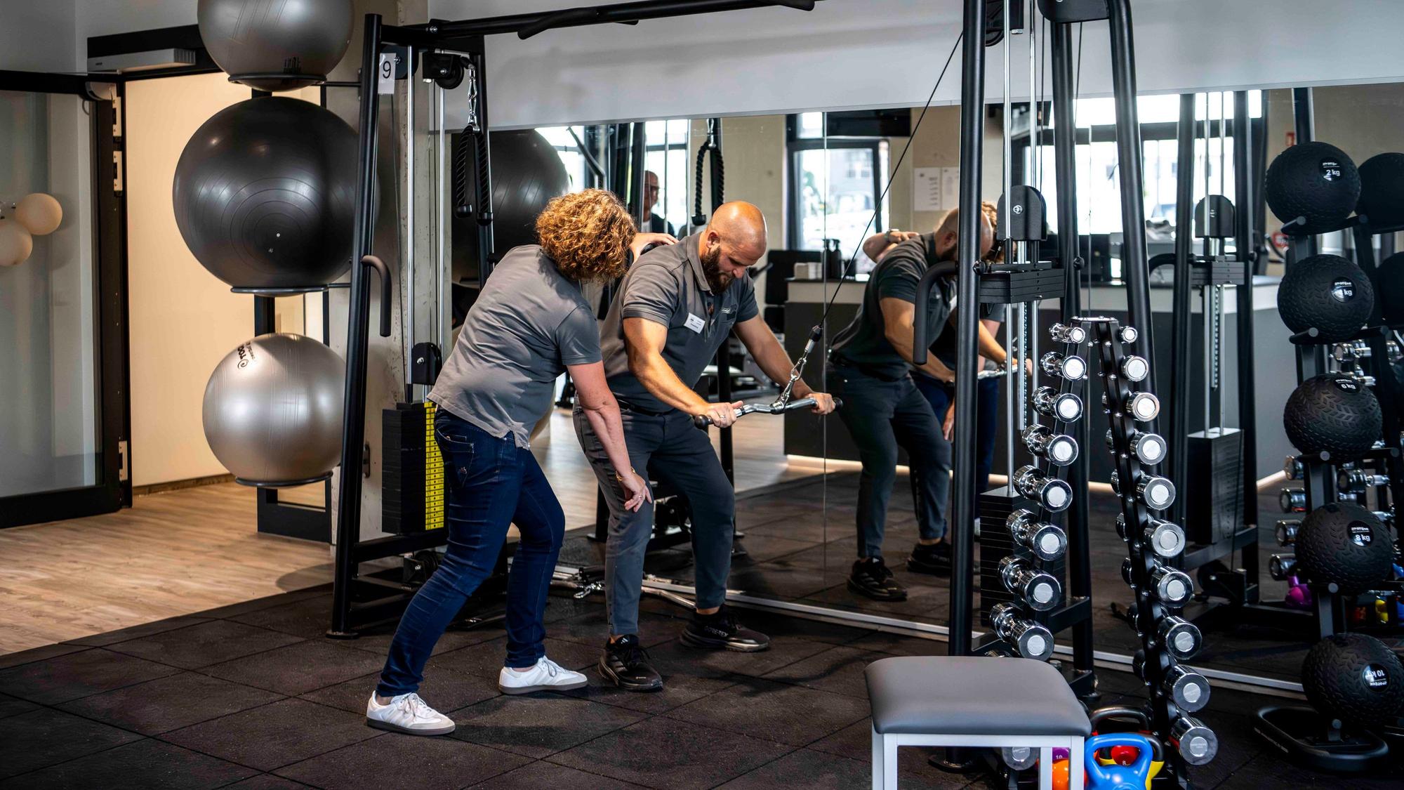 Das Bild zeigt das Fitness- und Kraftstudio der Physiotherapie-Spezialisten. Ein Mann macht eine Sportübung und wird dabei angeleitet.