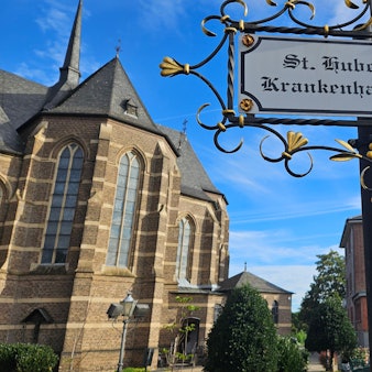 Zu sehen ist das Bedburger Krankenhaus neben der Kirche, im Vordergrund steht ein Schild, das auf das Krankenhaus verweist.