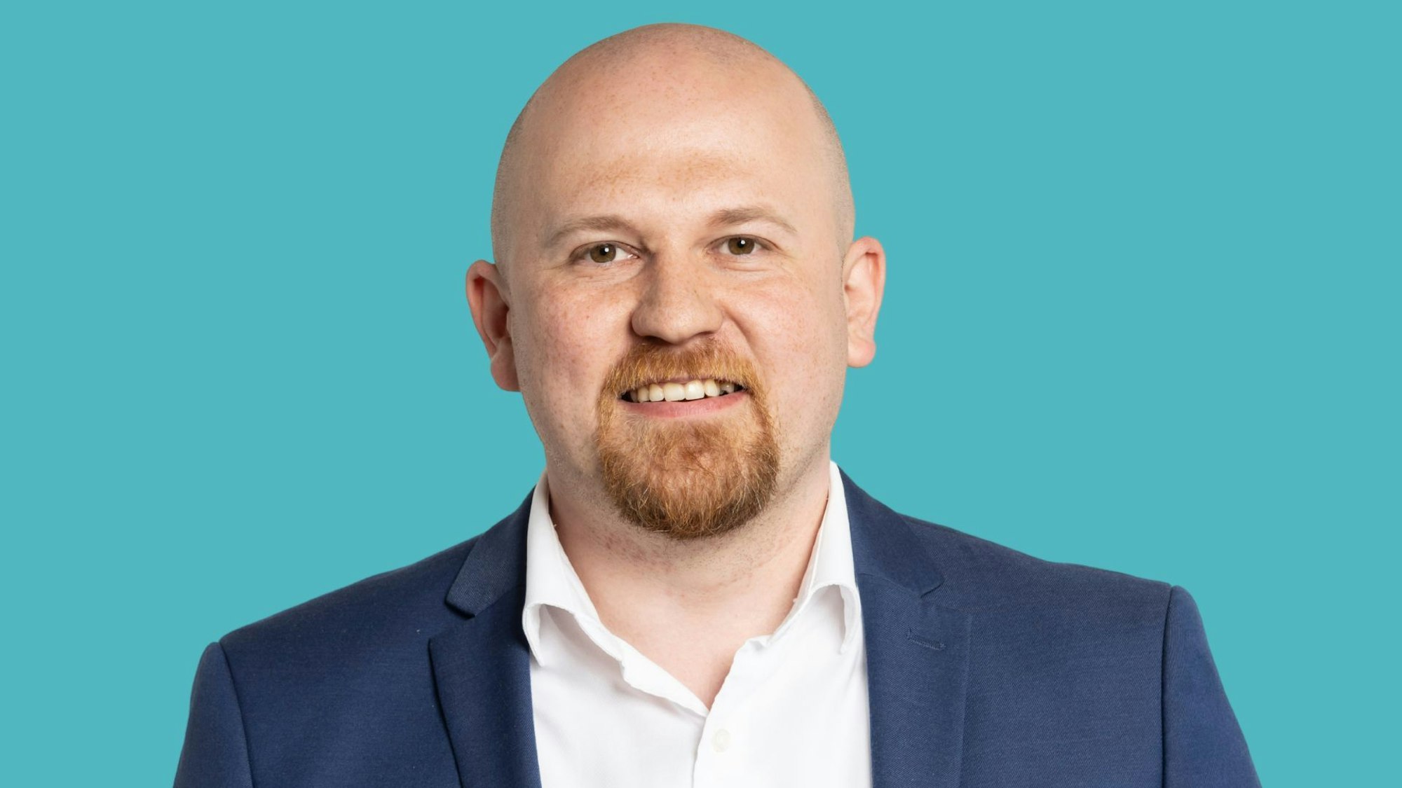 Martin Finder steht in blauem Sakko und weißem Hemd vor einem türkisen Hintergrund. Er trägt keine Krawatte.
