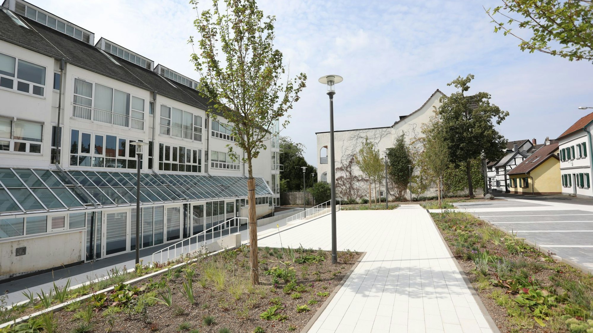 Beete und Bäume auf einem Platz mit einem hellen Gehweg, am Rande mehrere Parkplätze.