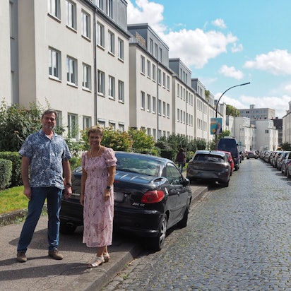Ein Mann und eine Frau stehen auf einem Bürgersteig, an dessen Straße viele Autos geparkt sind.