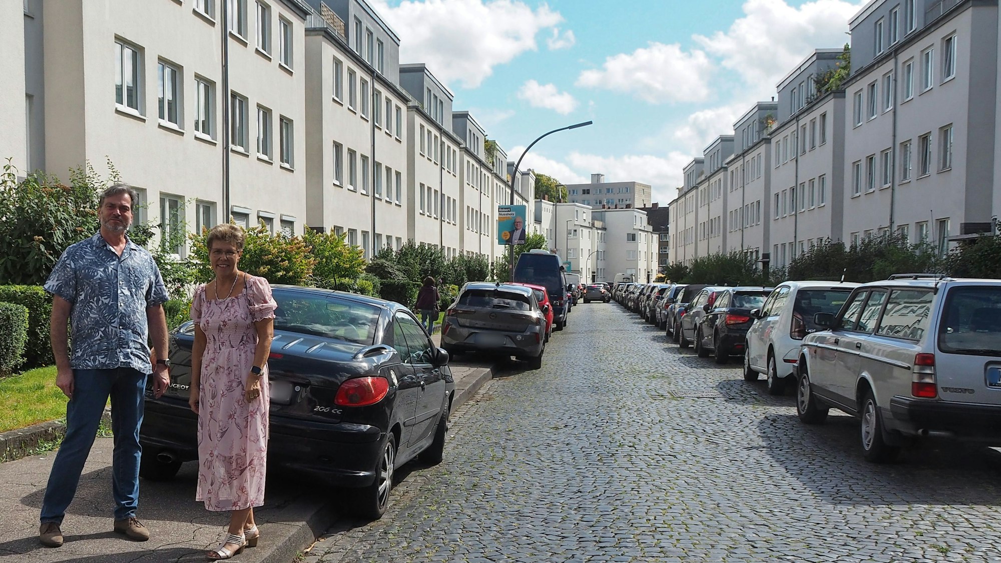 Ein Mann und eine Frau stehen auf einem Bürgersteig, an dessen Straße viele Autos geparkt sind.