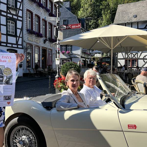 Eine Frau steht auf dem Marktplatz in Monschau und hält ein Plakat in den Händen. Daneben sitzen ein Mann und eine Frau in einem Austin-Healey-Oldtimer.