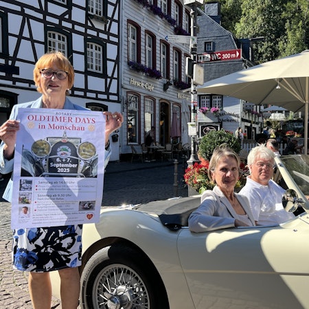 Eine Frau steht auf dem Marktplatz in Monschau und hält ein Plakat in den Händen. Daneben sitzen ein Mann und eine Frau in einem Austin-Healey-Oldtimer.