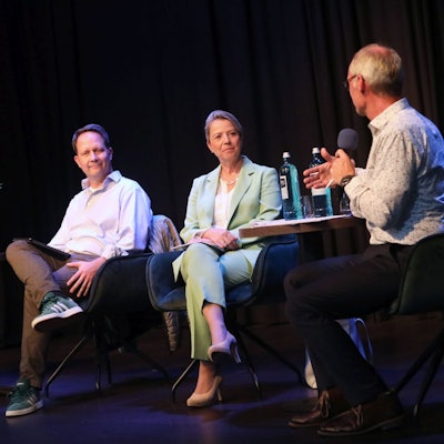 Drei Männer und eine Frau sitzen auf Sesseln, die auf einer Bühne stehen.