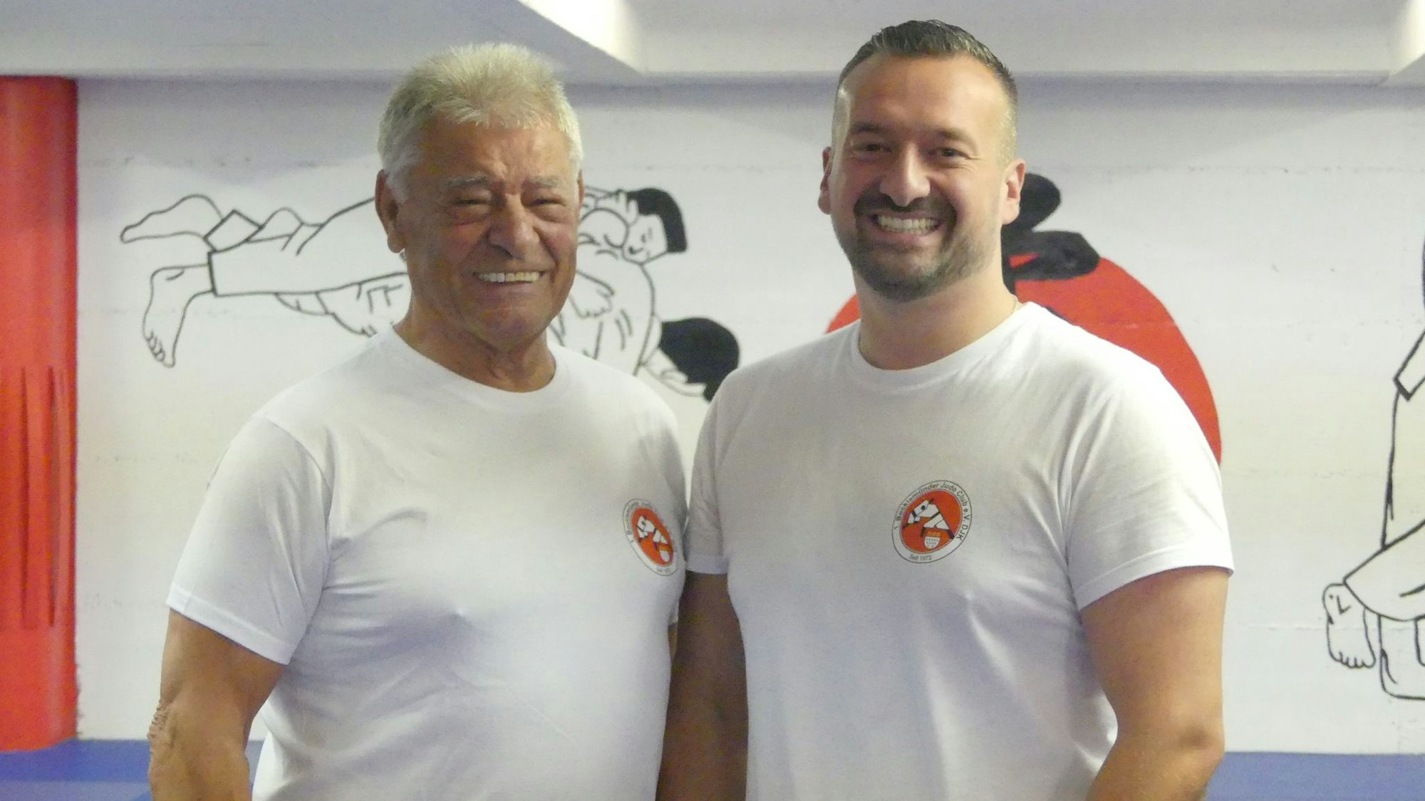 Ein grauhaariger Mann steht neben einem jüngeren Mann mit dunklem Haar in einem Trainingsraum mit blauem Bodenbelag.