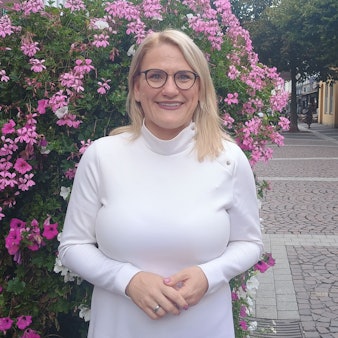 Christina Stumpp steht in der Brühler Innenstadt vor einem blumigen Busch. Sie trägt ein weißes Oberteil und eine Brille.