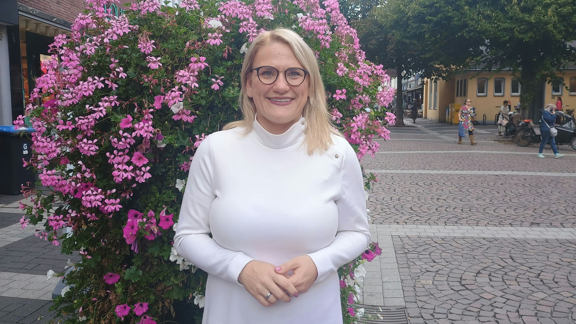 Christina Stumpp steht in der Brühler Innenstadt vor einem blumigen Busch. Sie trägt ein weißes Oberteil und eine Brille.
