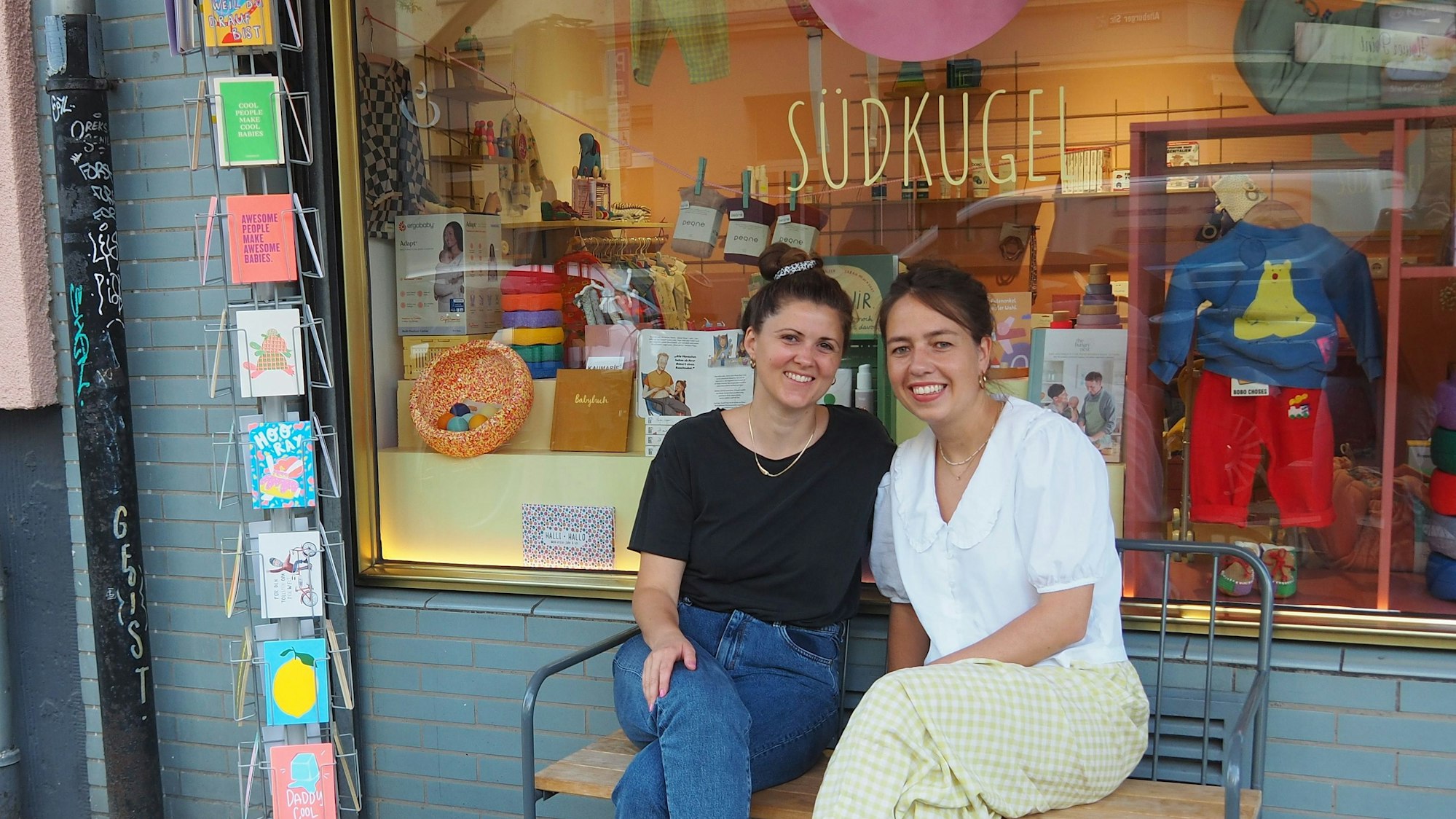 Zwei junge Frauen sitzen auf einer Bank vor dem Schaufenster eines Geschäfts mit Baby-Zubehör.