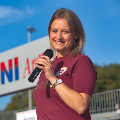 Eine junge Frau steht mit einem Mikrofon in der Hand vor einem Fußballplatz