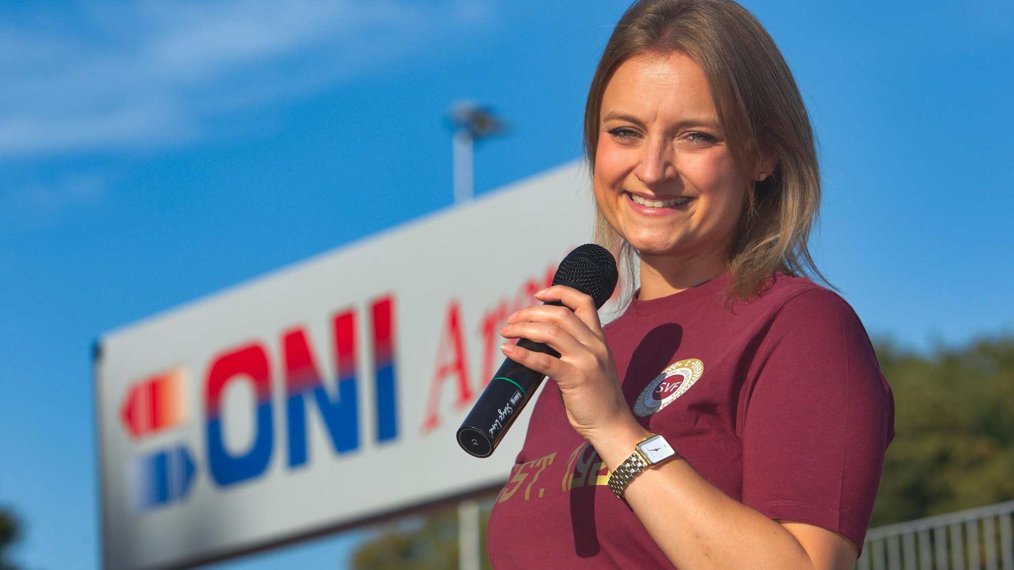 Eine junge Frau steht mit einem Mikrofon in der Hand vor einem Fußballplatz