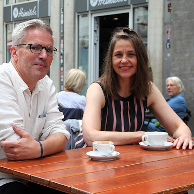 Ein Mann und eine Frau sitzen draußen in einem Café an einem Holztisch, auf dem zwei Kaffeetassen stehen.