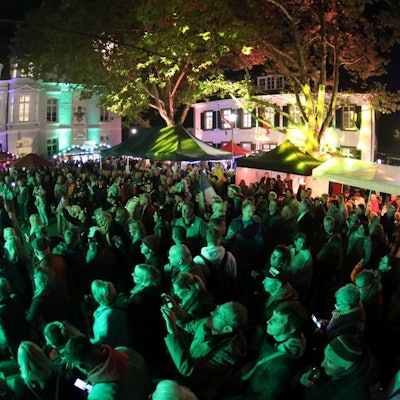 Menschen auf einem Marktplatz, alle in grünes Licht getaucht. Im Hintergrund das Rathaus Königswinter.