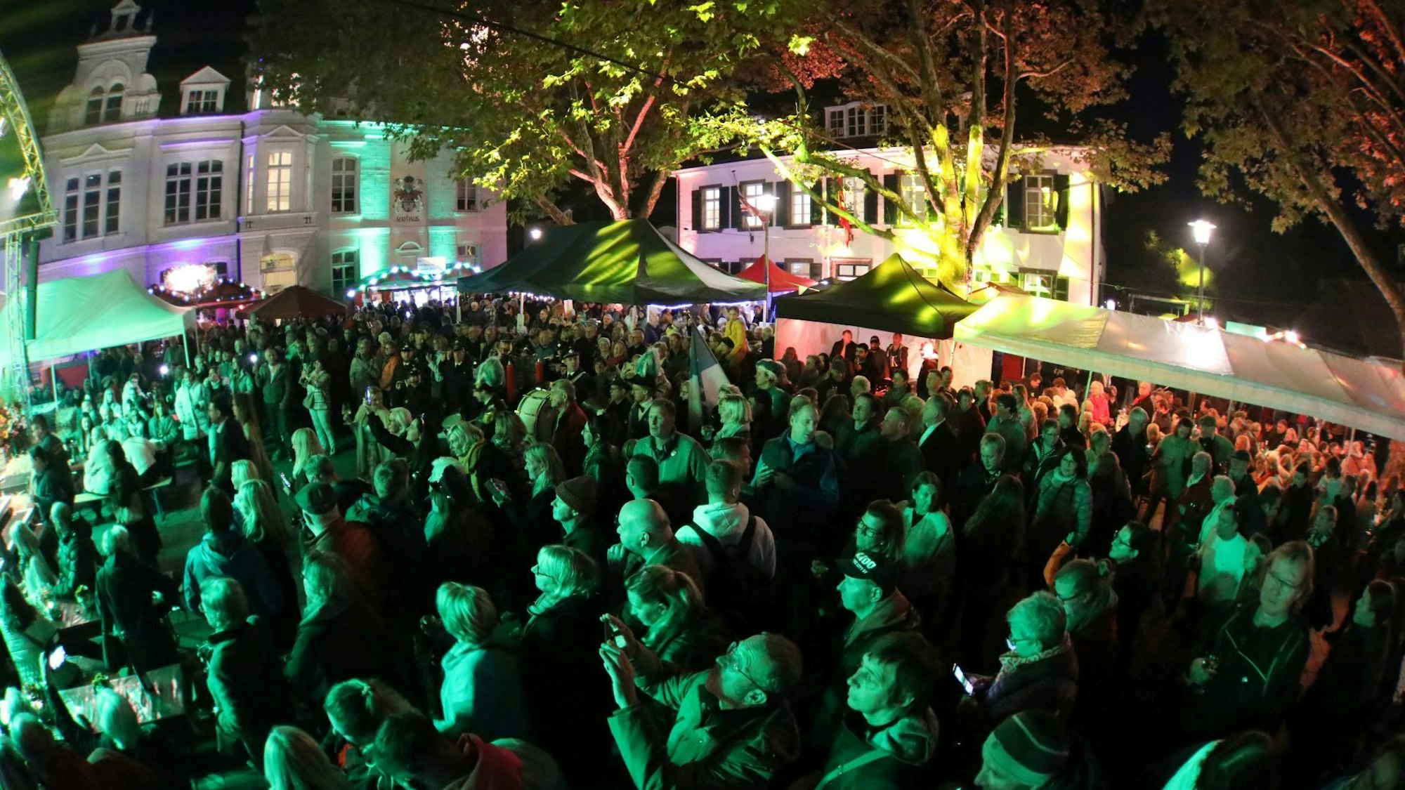 Menschen auf einem Marktplatz, alle in grünes Licht getaucht. Im Hintergrund das Rathaus Königswinter.