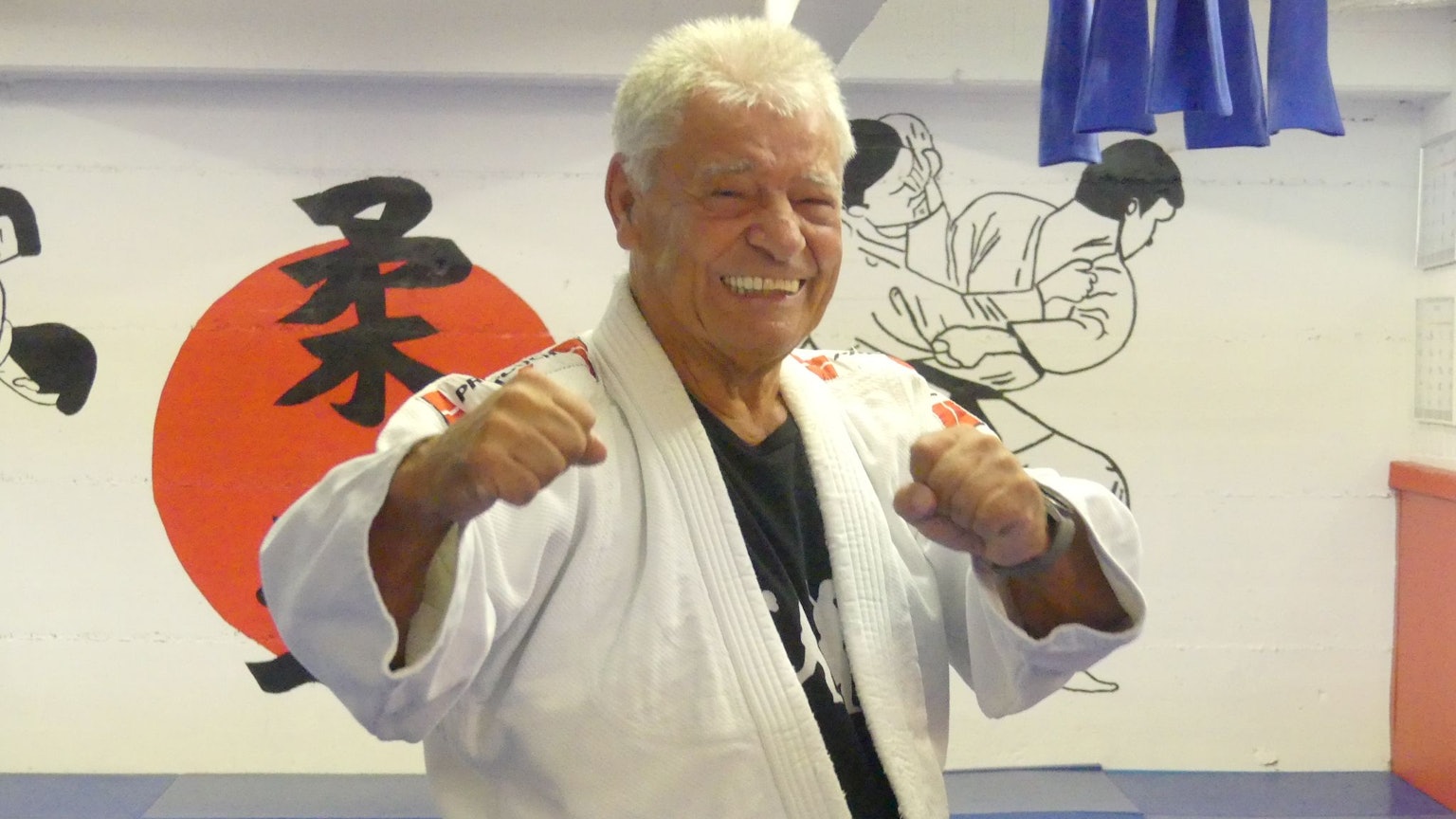 Ein Mann im Judo-Dress steht in einer Halle, die mit japanischen Zeichen und Bildern vom Judo geschmückt ist.