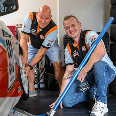 Reiner und Yannick Habeth grinsen neben einem Rennauto. Reiner Habeth fasst an den vorderen Reifen des Fahrzeugs. Yannick Habeth kniet daneben und hält Werkzeug.