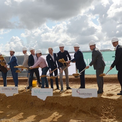 Elf Männer mit Schutzhelmen werfen, in einer Reihe stehend, mit Spaten Erde in die Luft.