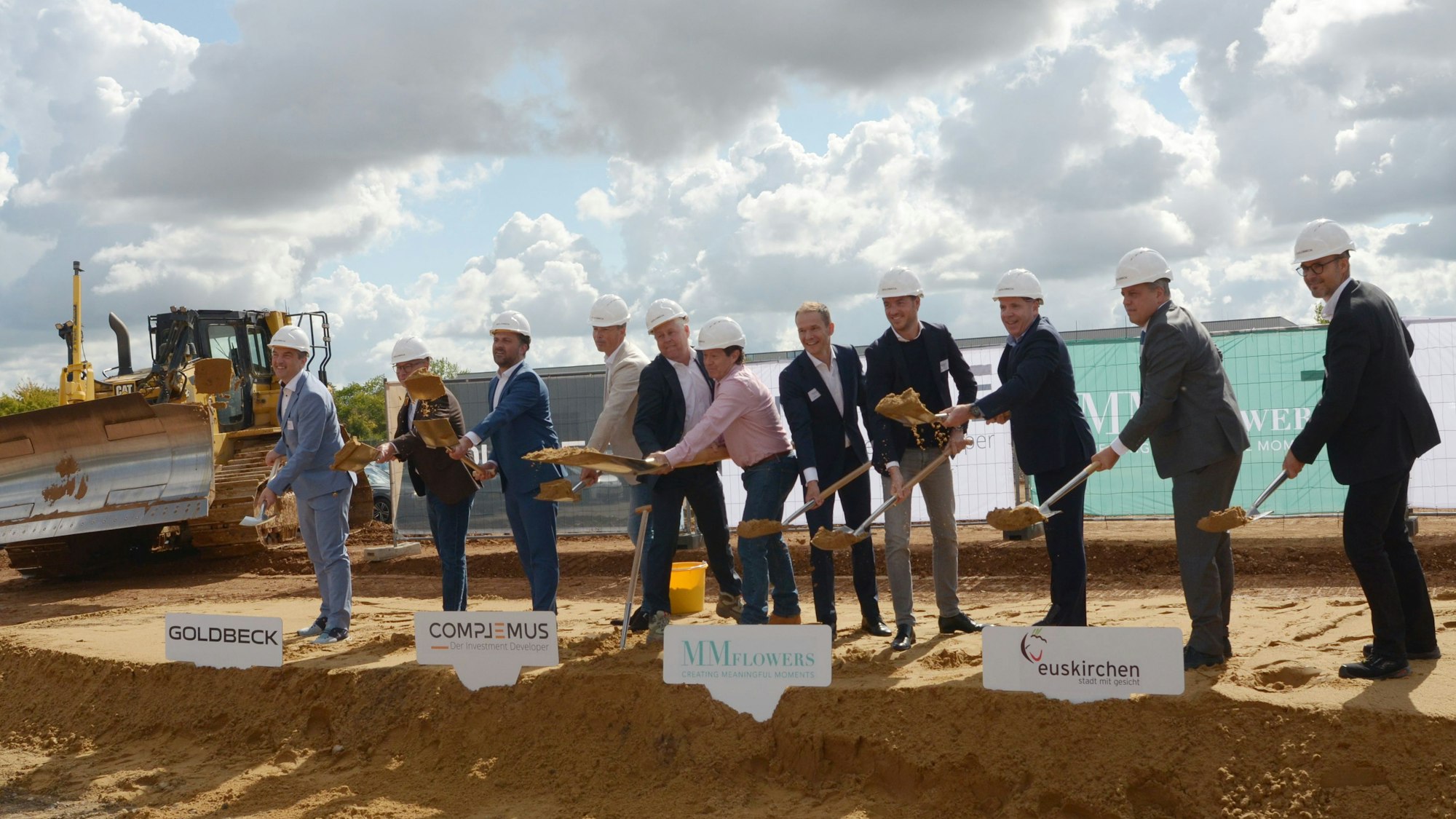 Elf Männer mit Schutzhelmen werfen, in einer Reihe stehend, mit Spaten Erde in die Luft.