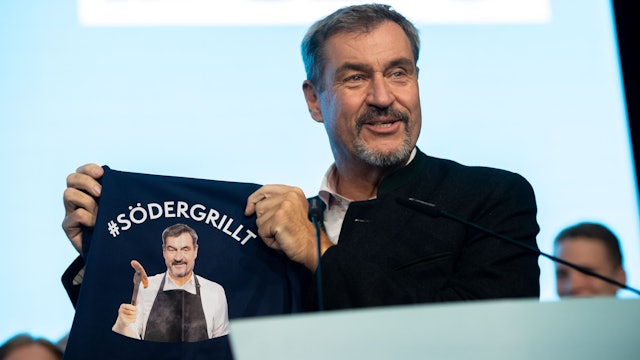Markus Söder (CSU) steht auf der Bühne und zeigt eine Grillschürze mit der Aufschrift „#SÖDERGRILLT“.