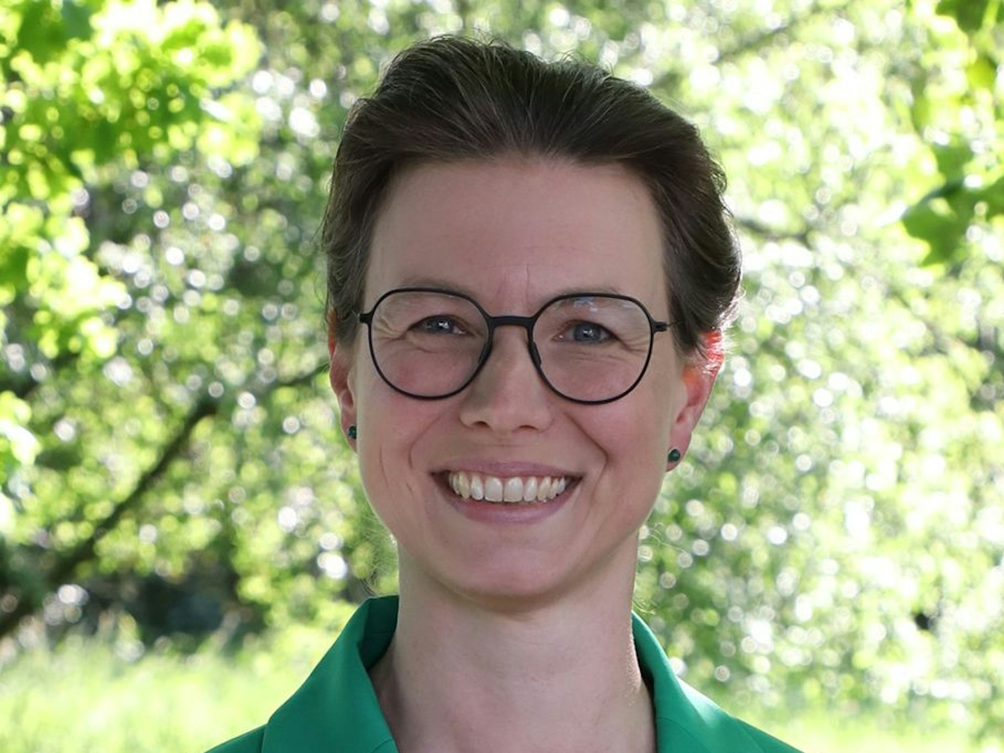 Dr. Simone Galliat trägt einen grünen Anzug und steht vor einem Baum.