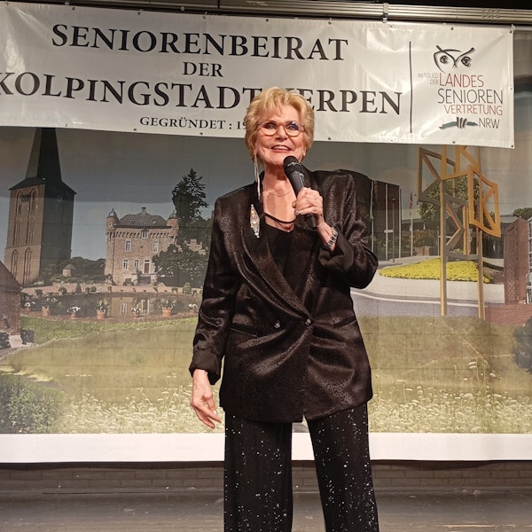 Peggy March steht mit einem Mikrofon auf der Bühne, sie hat kurze blonde Haare und trägt ein schwarzes Sakko.