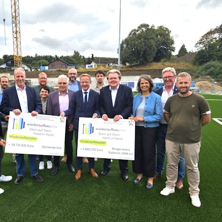 Eine Gruppe Menschen steht auf dem Kunstrasenplatz in Sötenich. Sie halten zwei Förderbescheide für die Gemeinde Kall und den Bürgerverein in den Händen.