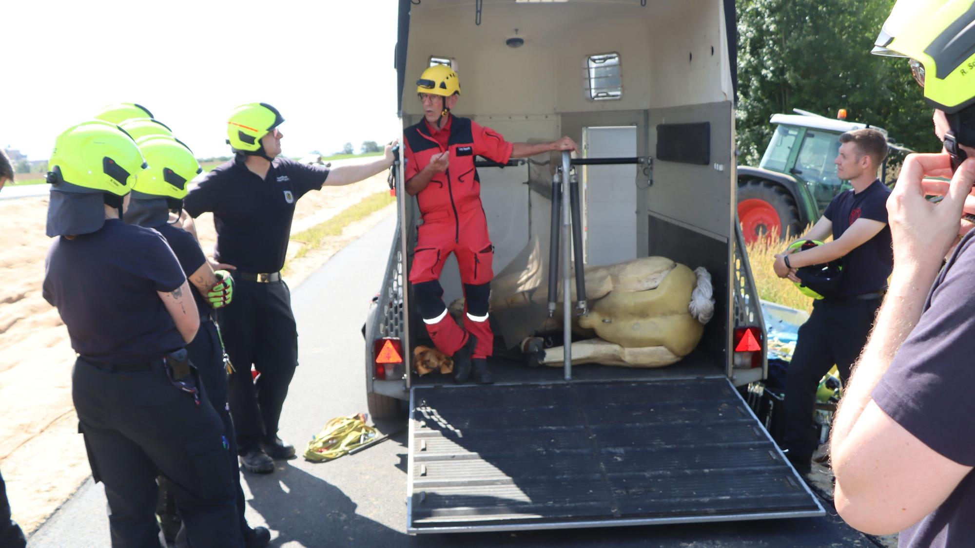 Auf dem Bild ist ein Pferdanhänger zu sehen, in dem der Pferde-Dummy auf der Seite liegt. Feuerwehrleute und der Seminarleiter stehen drumherum.