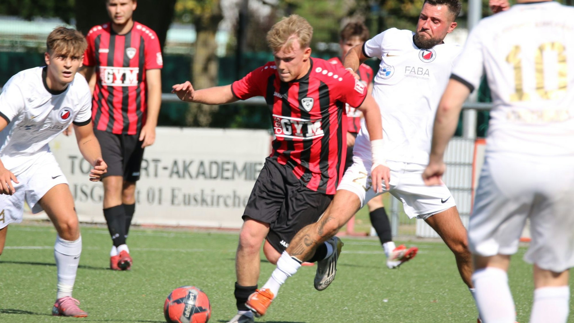 Das Bild zeigt einen Zweikampf zweier Spieler um den Ball. Weitere Akteure schauen zu.