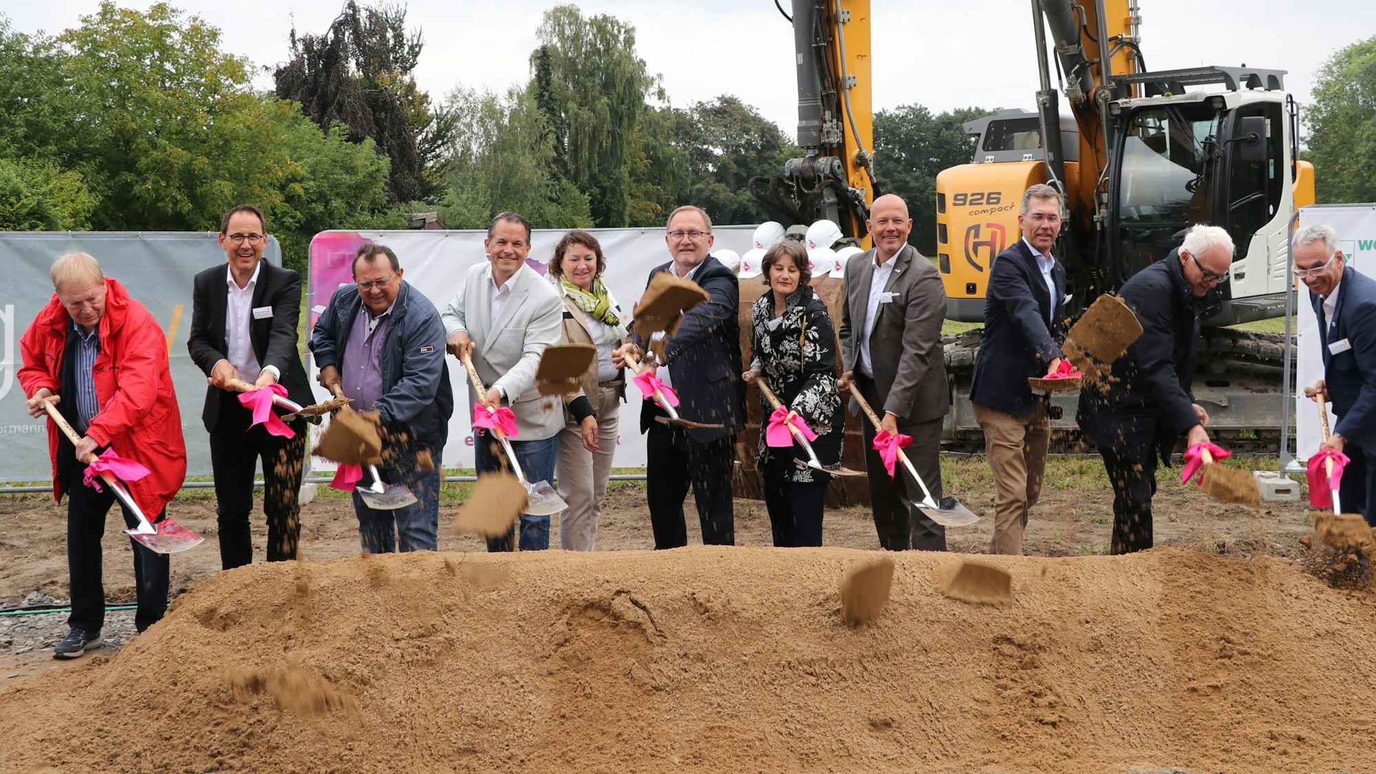 Auf dem Bild sind Männer und Frauen auf einer Baustelle zu sehen, die mit Spaten Sand in die Luft werfen - die Baustelle ist damit offiziell eröffnet.