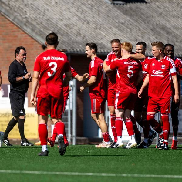 Zülpicher Spieler jubeln nach dem frühen 1:0. Ein Spieler am linken Bildrand hält sich ungläubig die Hand vors Gesicht. Der Schiedsrichter spricht mit den Spielern.