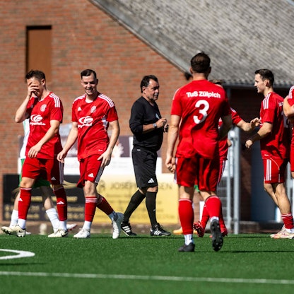 Zülpicher Spieler jubeln nach dem frühen 1:0. Ein Spieler am linken Bildrand hält sich ungläubig die Hand vors Gesicht. Der Schiedsrichter spricht mit den Spielern.