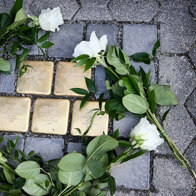 Das Bild zeigt fünf Pflastersteine aus Messing, auf denen Namen stehen. Drumherum liegen weiße Rosen.