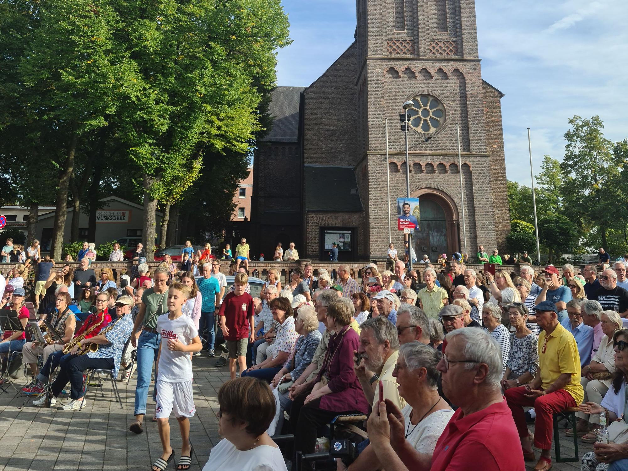 Unter einem Kirchturm sitzen viele Menschen auf einem Platz.