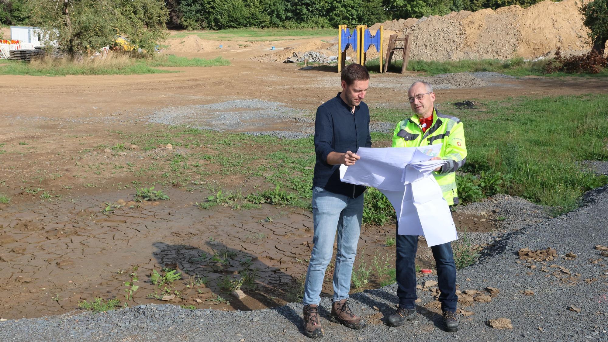 Bauamtsleiter Jan Foerster und Jörn Renke vom Planungsbüro Schumacher stehen auf einem freien Gelände und schauen auf einen Plan.